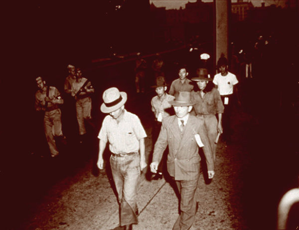 Double file line of Japanese Peruvian men walking, followed by armed MPs, New Orleans, Louisiana, ca. 1943